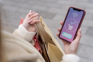 Mulher fazendo compra pelo celular e segurando uma sacola em uma de suas mãos para representar a compra